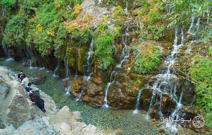آبشار «هفت‌چشمه» کرج؛ تعدادی آبشار در دل کوهی سرسبز جاری‌ست و چند نفر روی لبه‌ی صخره‌ی روبه‌روی آبشارها درحال راه‌رفتن‌اند.